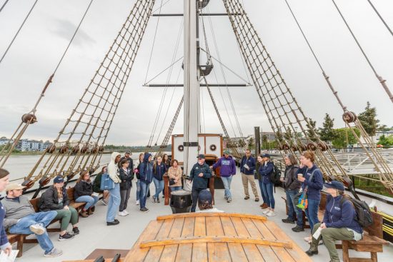 A tour group listens to a presentation on the deck of an old sailing ship.