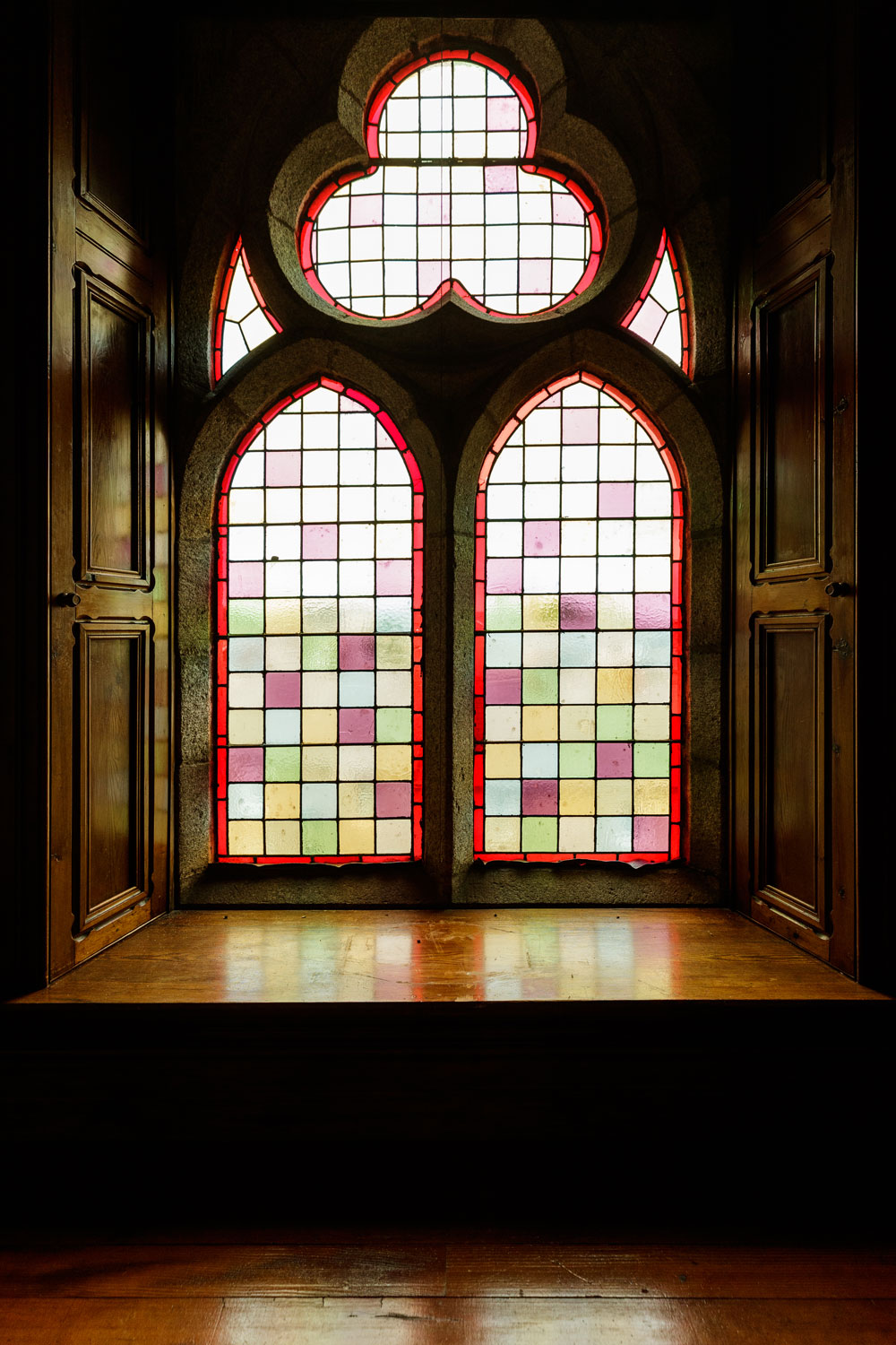 A stained glass window with uniquely-shaped panels flanked by wooden shutters and a large window seat.